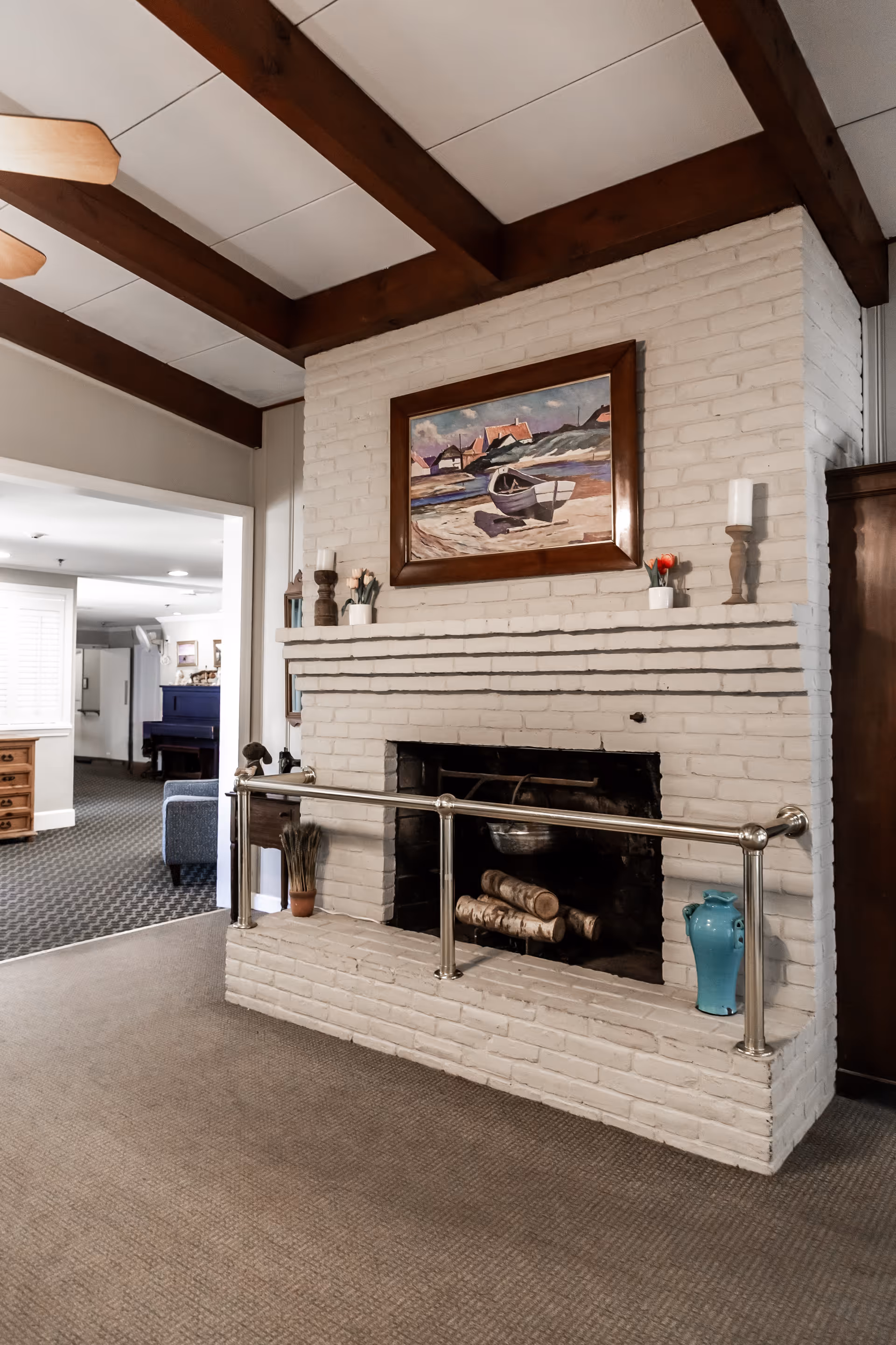 White brick fireplace with a framed painting above, decorative vases and candles, wood ceiling beams and a view into a lounge area.