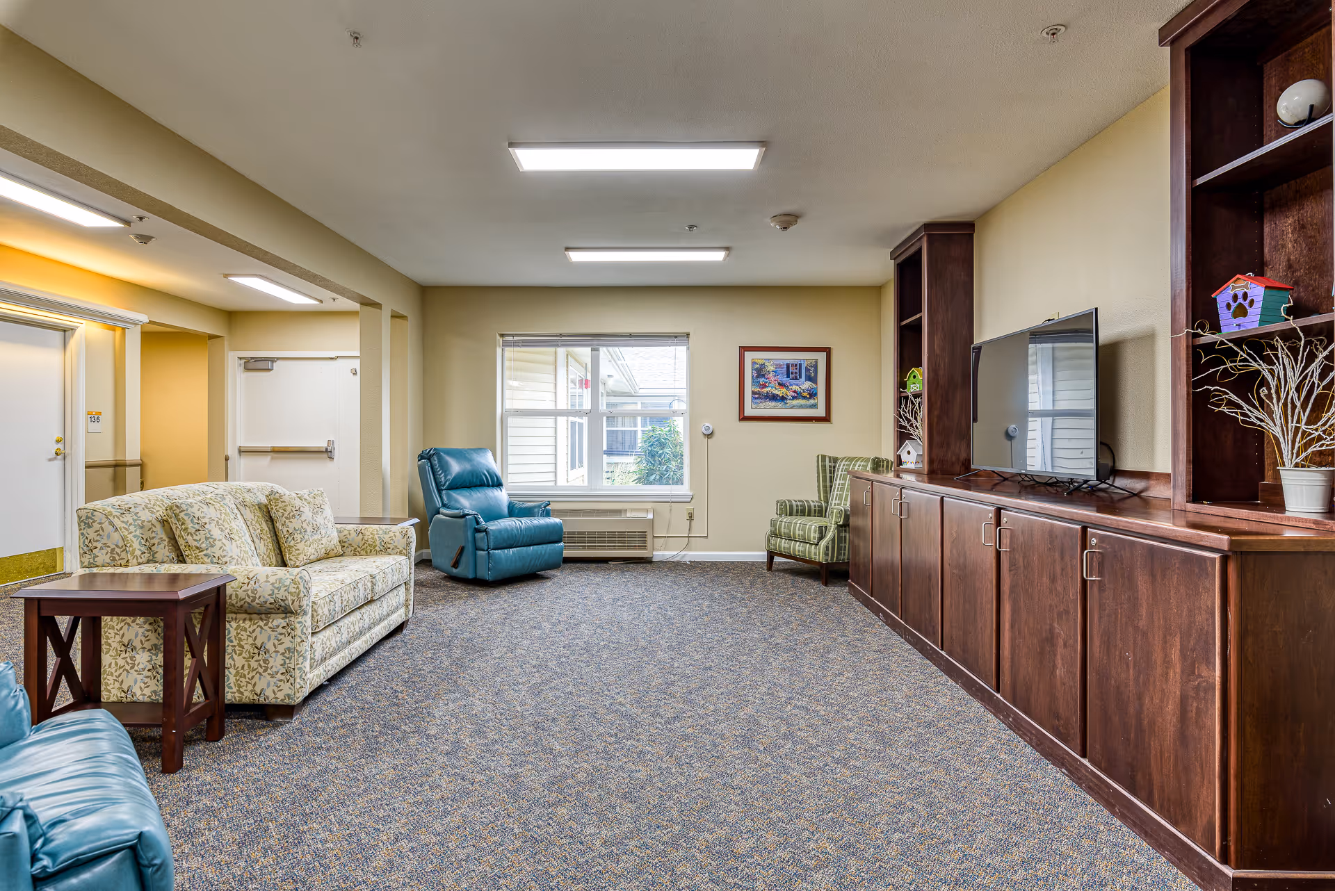 A spacious living room area in an assisted living facility with a floral patterned sofa, a blue recliner chair, a green plaid armchair, and a large wooden entertainment center with a flat-screen TV and decorative items. The room has beige walls, a window letting in natural light, and carpeted flooring.