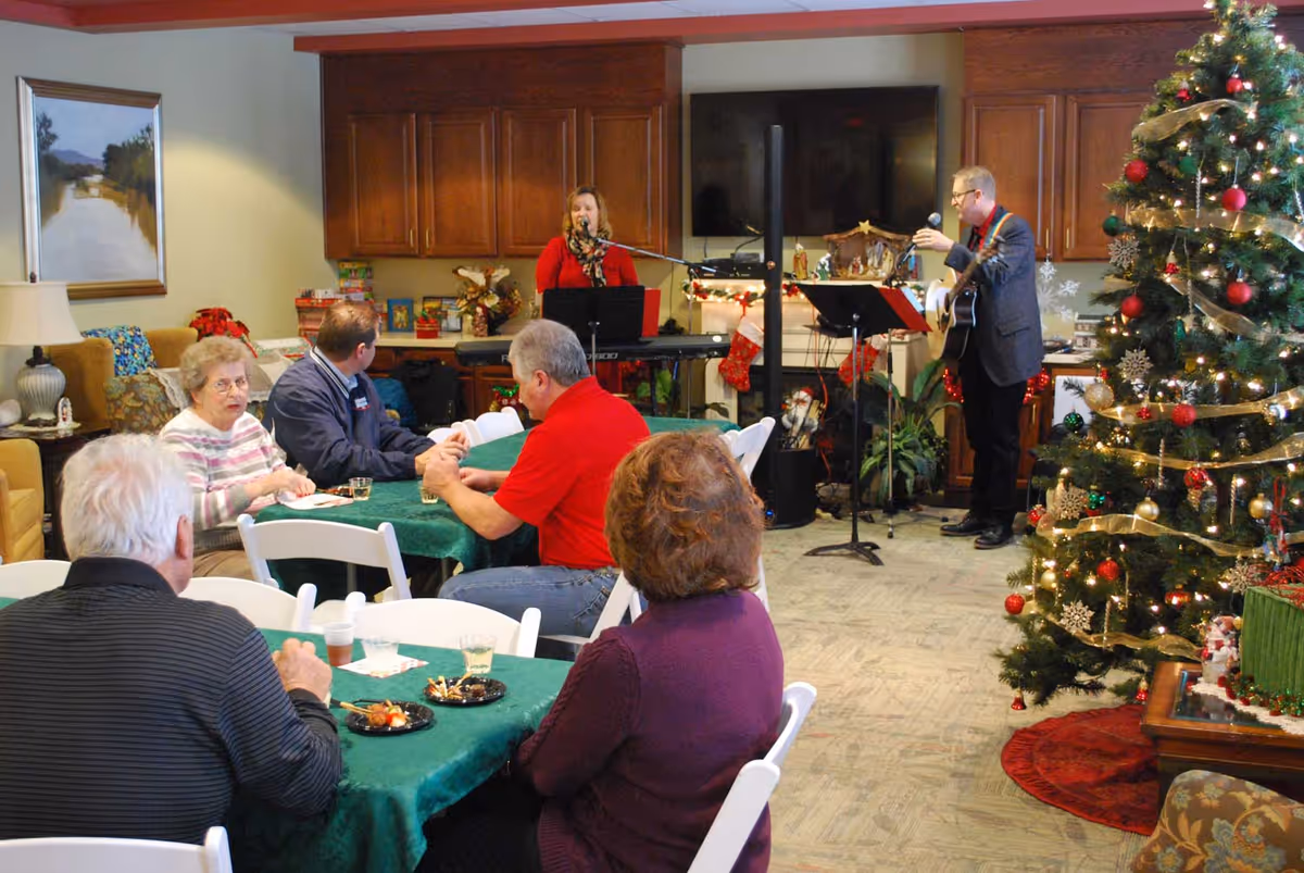 A group of elderly people seated at tables with green tablecloths in a common room decorated for Christmas. Two musicians, a woman playing keyboard and a man playing guitar, are performing near a large decorated Christmas tree. The room has wooden cabinets, a TV, and festive decorations including stockings and a nativity scene.