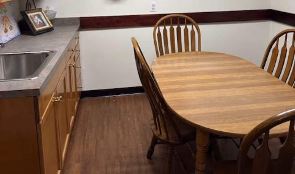 Wood dining table and chairs beside a kitchenette in a senior living facility.