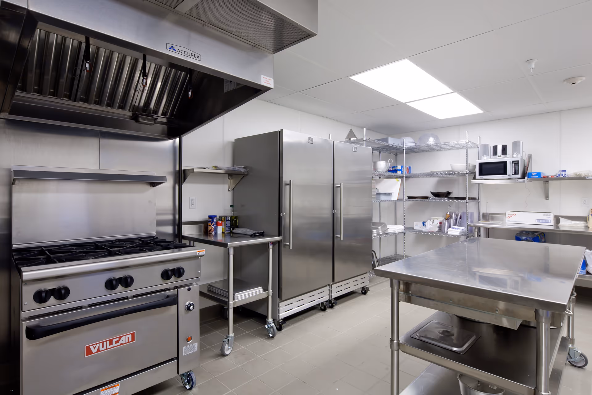A clean commercial kitchen with stainless steel appliances including a Vulcan stove with an overhead vent, two large refrigerators, metal shelving stocked with kitchen supplies, a microwave, and a stainless steel work table in the center.