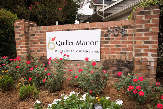 Brick entrance sign reading "Quillen Manor Independent & Assisted Living" with the number 709 and flower beds in front.