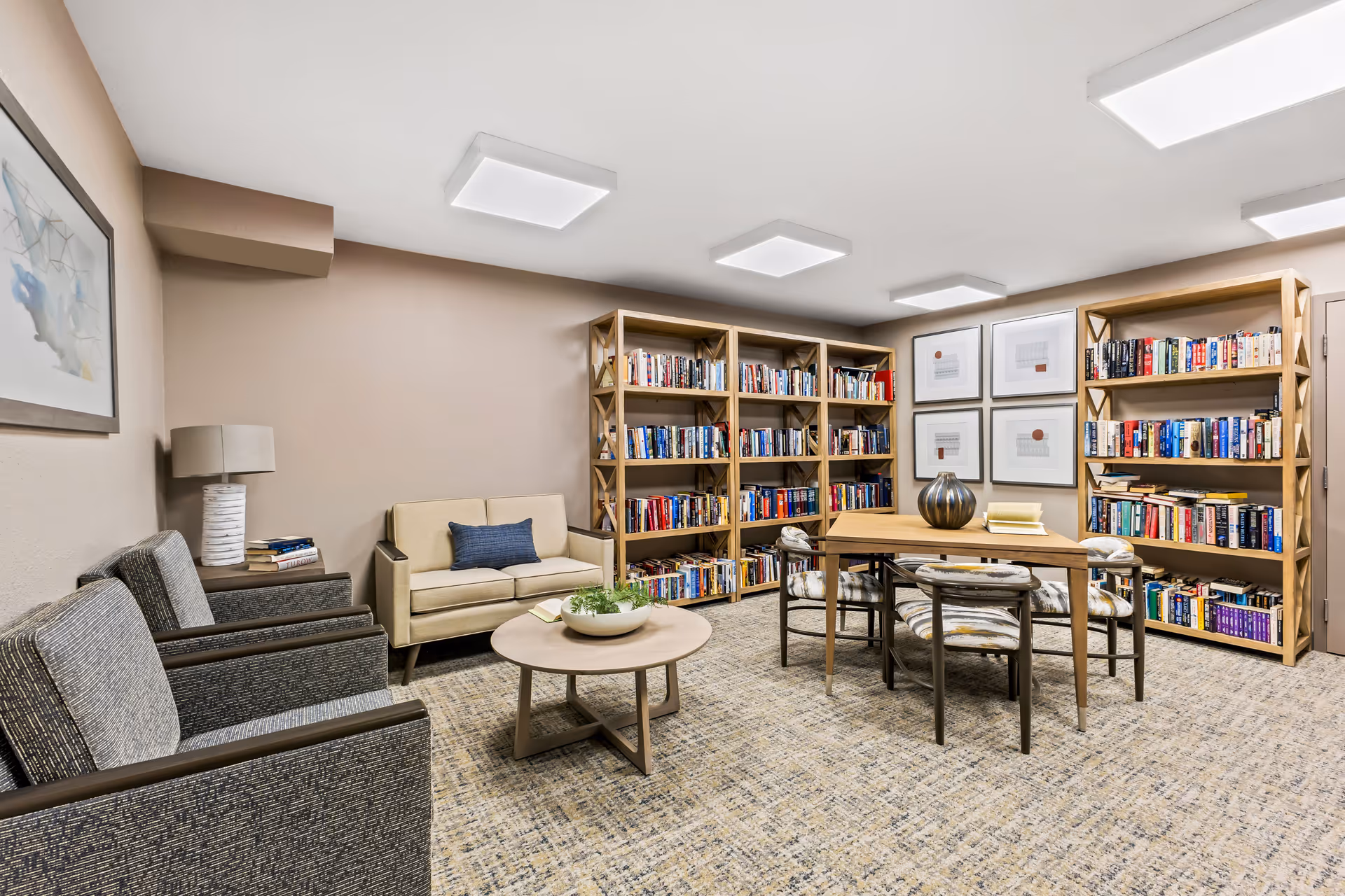 A cozy reading room with beige walls and carpeted floor featuring two gray armchairs and a beige loveseat with a blue cushion. There is a round wooden coffee table with a plant on it in front of the seating. The room has three wooden bookshelves filled with books and a wooden table with four chairs in the center. Four framed artworks hang on the wall above the bookshelves. The ceiling has modern square light fixtures.