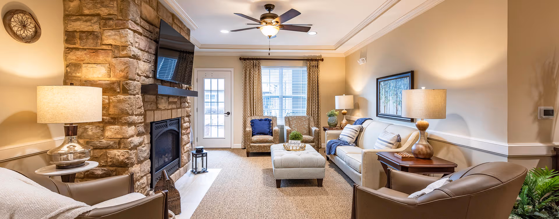 A cozy living room with a stone fireplace on the left wall, a mounted flat-screen TV above it, and a ceiling fan with light. The room features a beige sofa with pillows, two armchairs, a cushioned ottoman coffee table, side tables with lamps, a framed artwork on the wall, and a door with windows leading outside. The room is warmly lit and decorated with neutral tones and some greenery.