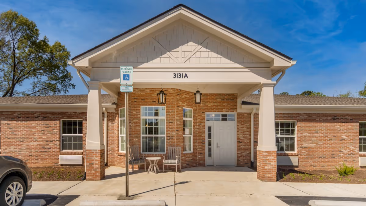 Front entrance of a single-story brick building with a covered porch supported by columns, two chairs and a small table on the porch, a reserved parking sign for handicapped and van accessible parking, and a clear blue sky above.