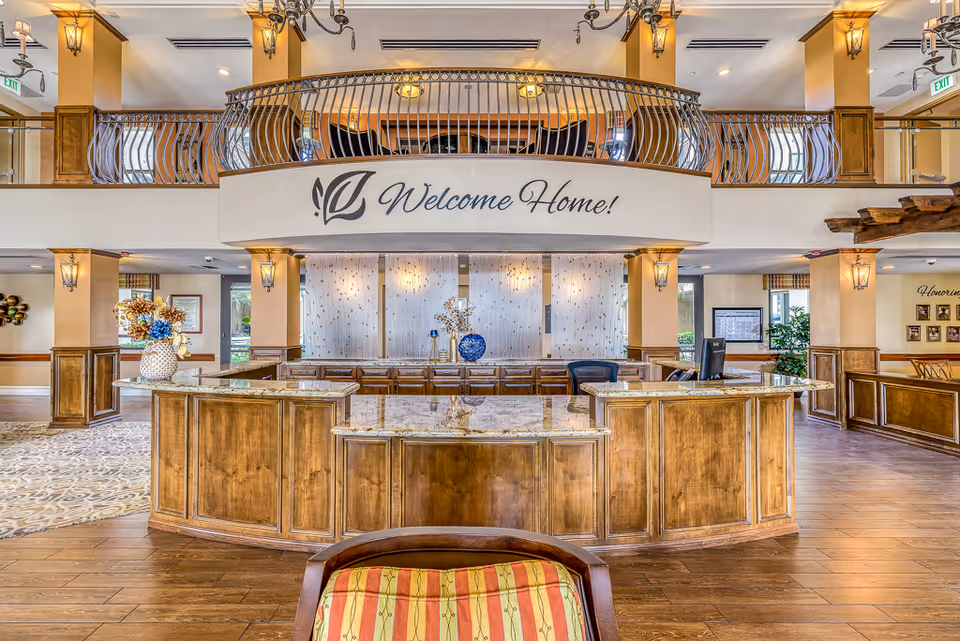 Spacious and warmly lit reception area with a large wooden front desk topped with granite. Behind the desk is a decorative wall with soft lighting and a sign above that reads 'Welcome Home!'. The area features wooden paneling, elegant light fixtures, and a balcony railing above. There are decorative vases with flowers on the desk and a comfortable chair with a striped cushion in the foreground.