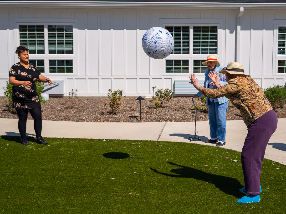 Three women playing with a large inflatable ball on a grassy area outside a white building with large windows. One woman is about to catch the ball, another woman stands ready, and the third woman uses a walker and watches the game.
