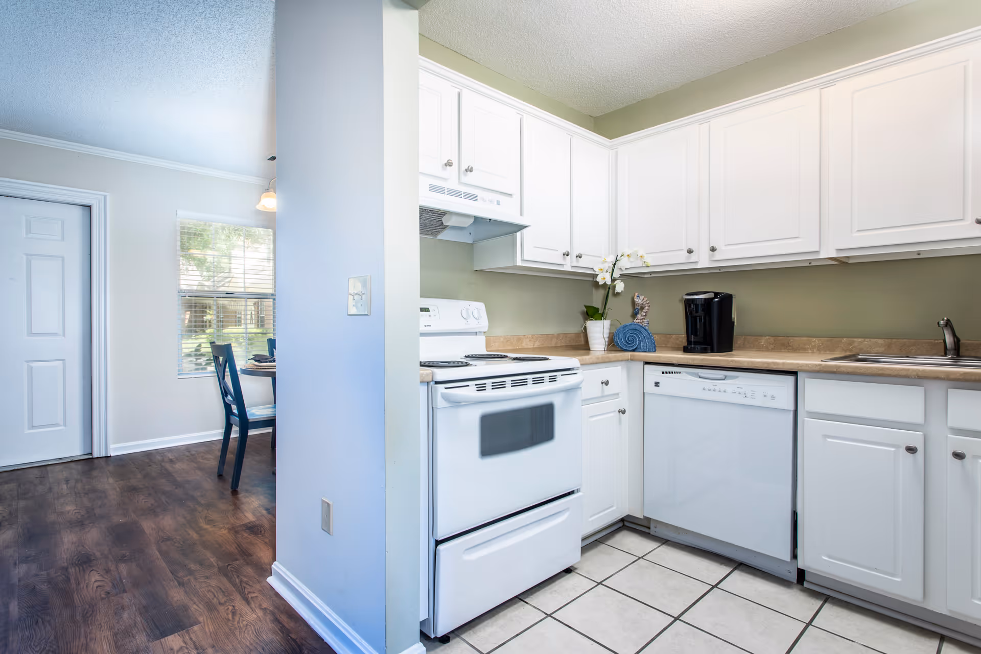 Bright kitchen with white cabinets, a white stove, dishwasher, and a coffee maker on the countertop. The kitchen has a tiled floor and a greenish wall. Adjacent to the kitchen is a dining area with a window, wooden floor, and a table with chairs.