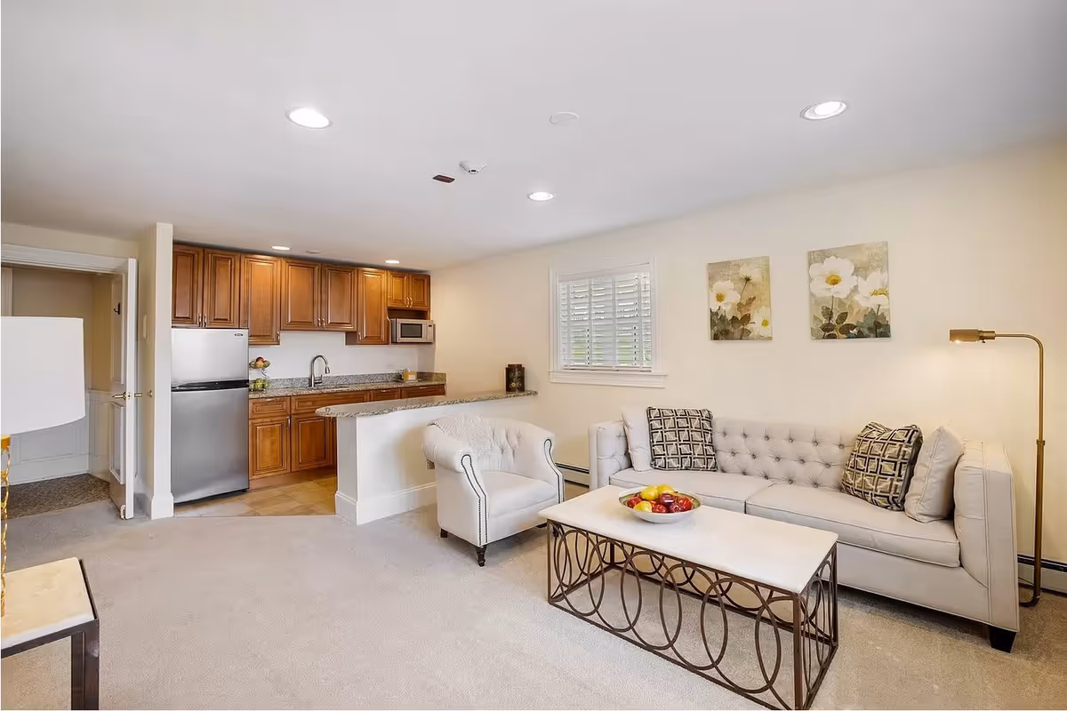 A cozy living area with a beige tufted sofa, an armchair, and a coffee table with a bowl of fruit. The room has light-colored walls with two floral paintings and a window with white shutters. In the background, there is a small kitchen with wooden cabinets, a stainless steel refrigerator, microwave, and granite countertops.