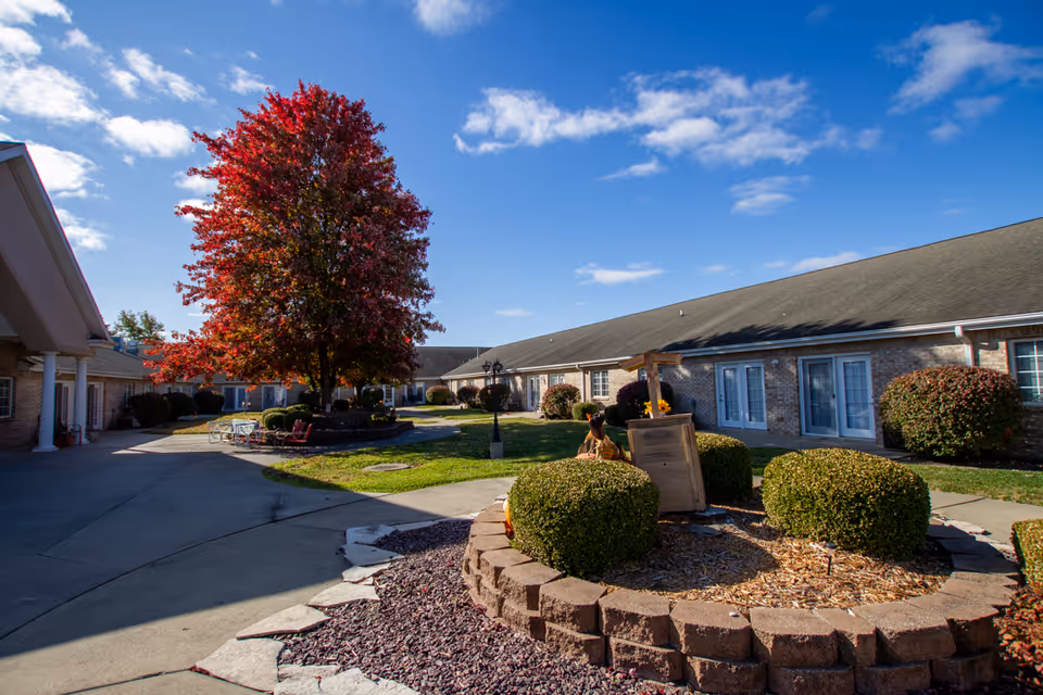 Outdoor courtyard area of a senior living facility with a large tree with red leaves in the center, surrounded by bushes and a circular stone planter. The courtyard is bordered by single-story brick buildings with multiple doors and windows. The sky is blue with some clouds.