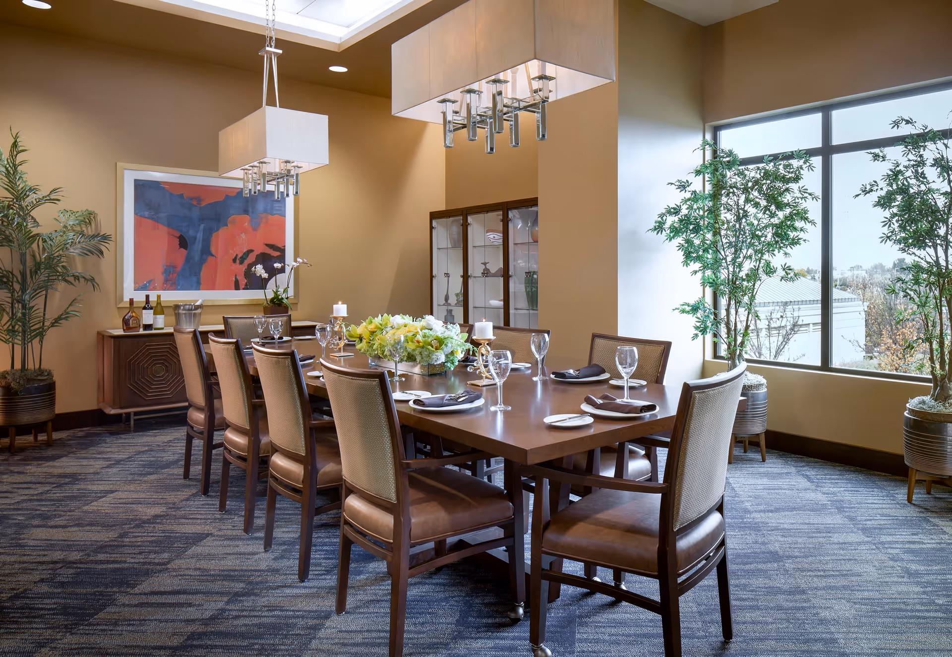A well-lit dining room with a long wooden table set for eight people, featuring wine glasses, plates, napkins, and a floral centerpiece. The room has large windows with natural light, potted plants, modern hanging light fixtures, a sideboard with bottles and glasses, and a colorful abstract painting on the wall.