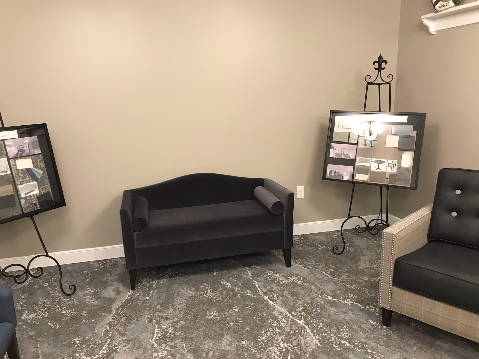A small seating area with a dark gray loveseat and a beige armchair with button tufting. Two black metal easels hold framed boards with various fabric and design samples. The floor has a gray marbled pattern, and the walls are painted beige.