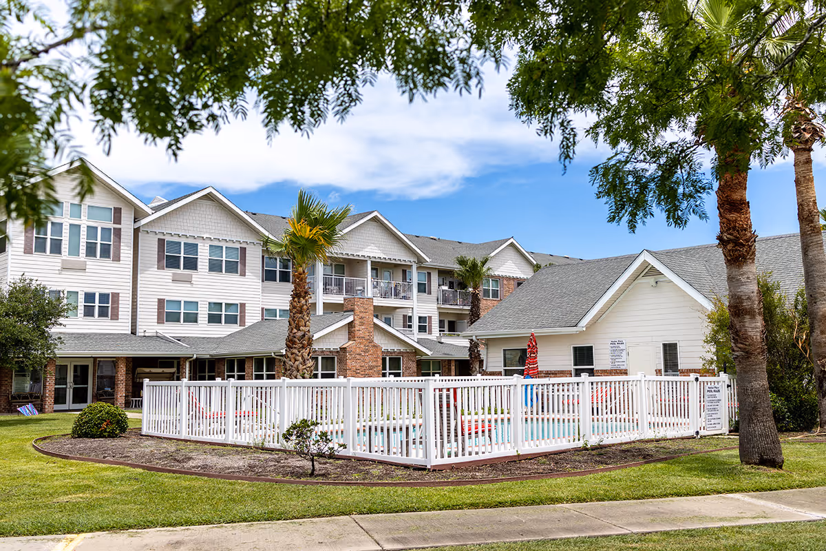 Exterior view of a senior living facility with a white three-story building, palm trees, and a fenced swimming pool area under a partly cloudy sky.