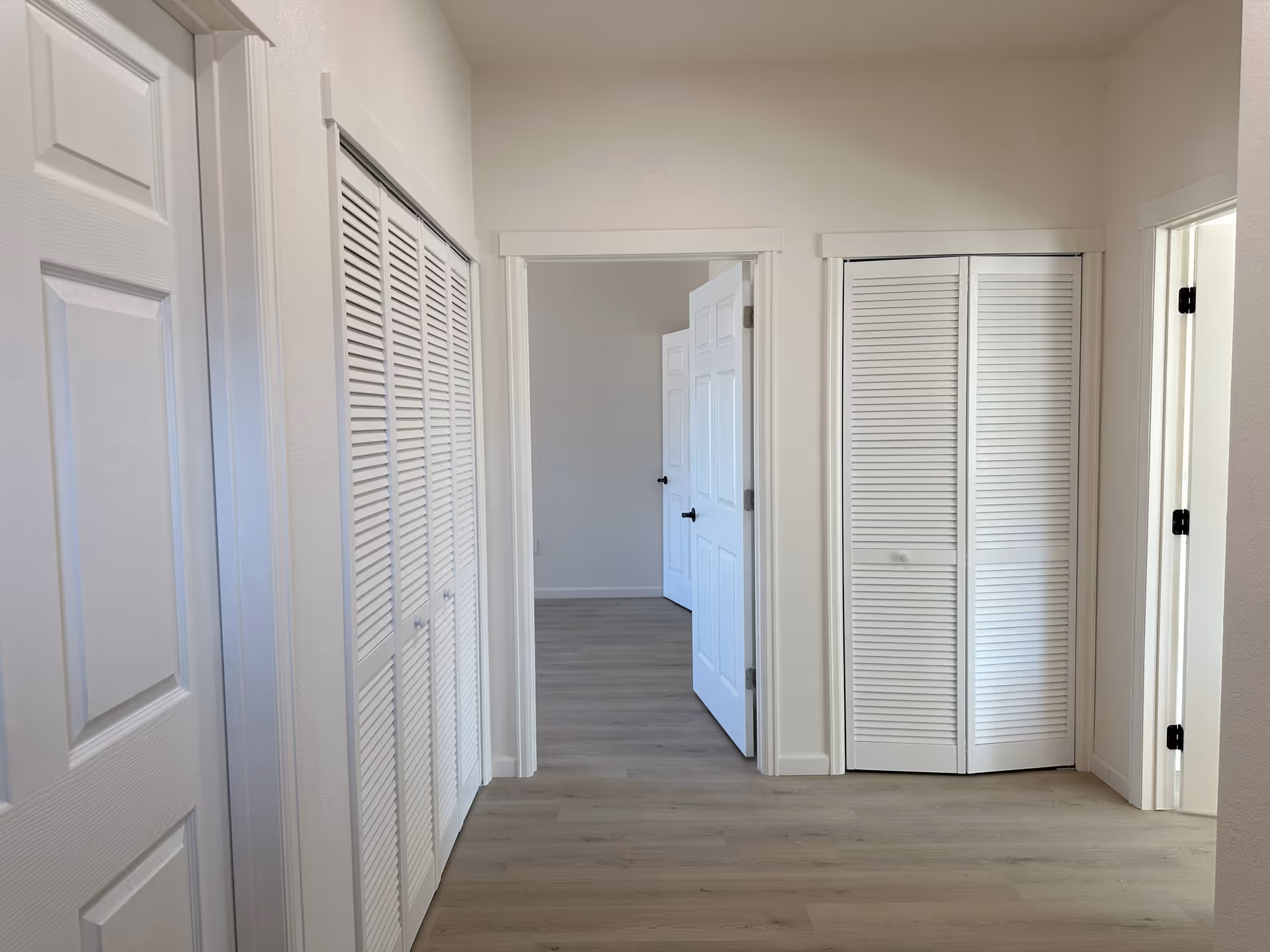 Interior hallway with light wood flooring and white walls. There are multiple white doors, including louvered closet doors on both sides and a partially open door leading to another room.