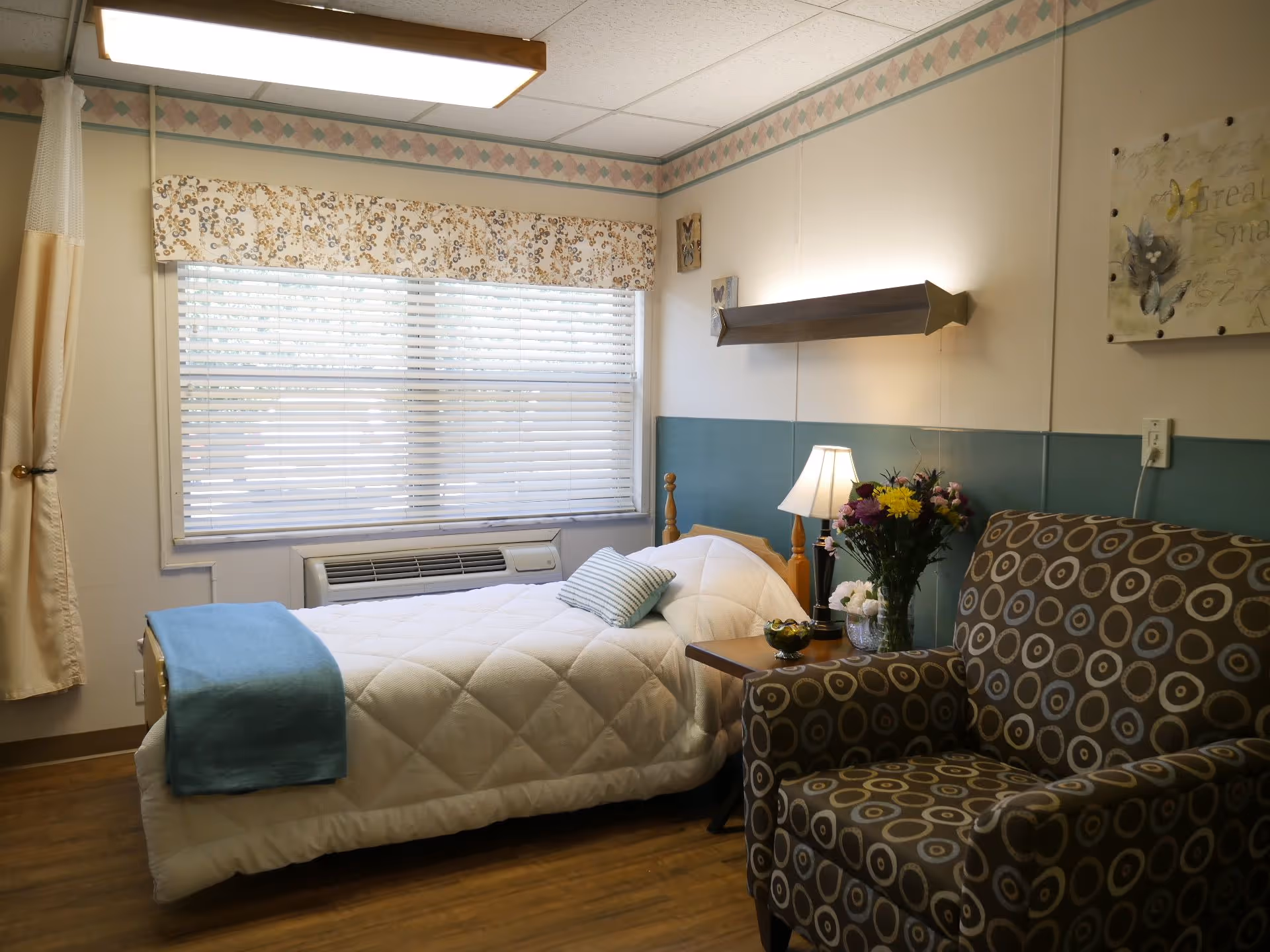 A cozy bedroom in a senior living facility with a single bed covered in a white quilt and a blue throw blanket. There is a patterned armchair with circular designs next to a small wooden side table holding a lamp and a vase of colorful flowers. A large window with blinds and a floral valance lets in natural light. The walls have a decorative border near the ceiling and a teal panel behind the bed.