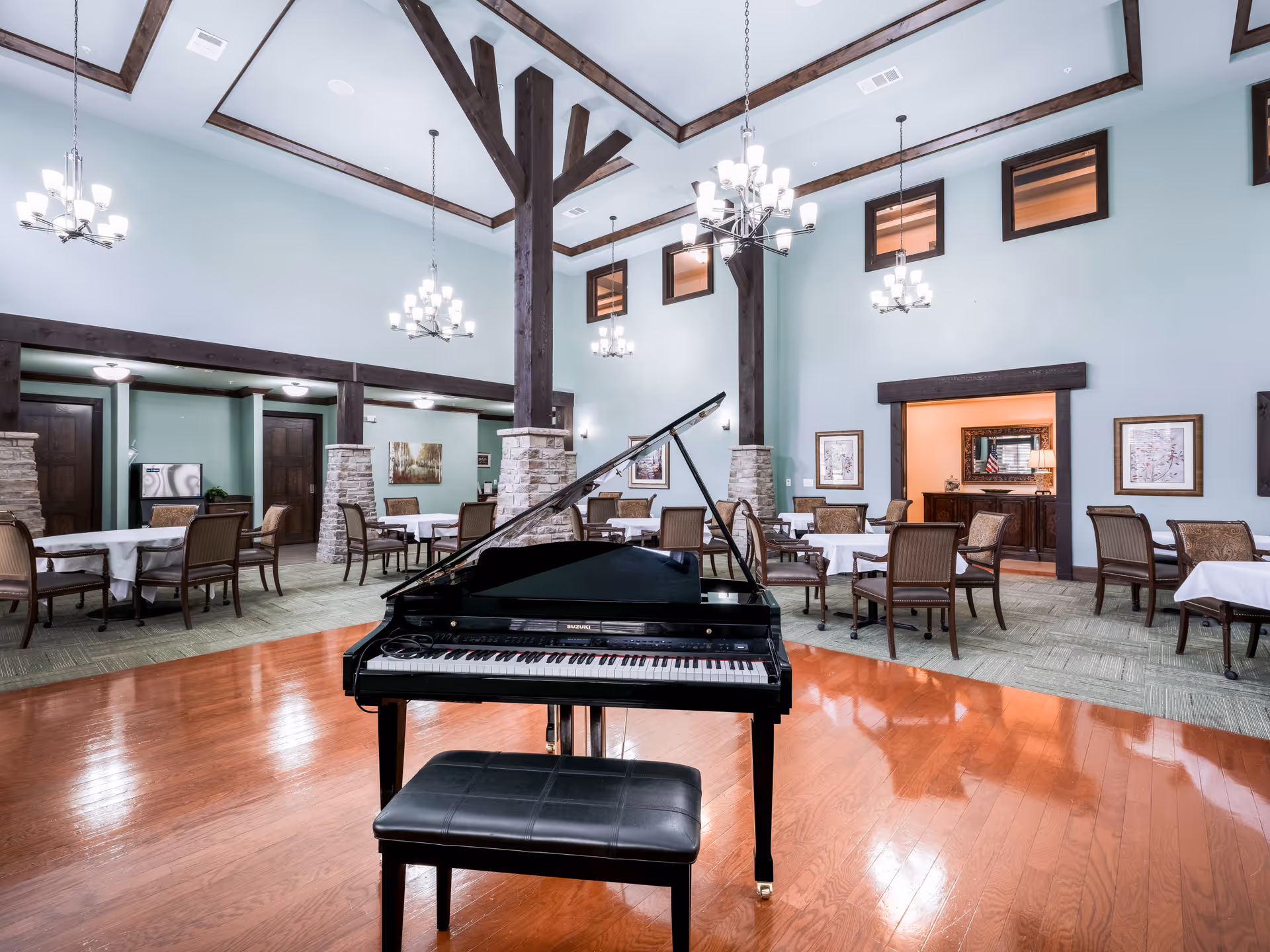 Spacious dining room with high ceilings, wooden beams, and multiple chandeliers. A black grand piano with a matching bench is positioned on a polished wooden floor in the foreground. Several tables with white tablecloths and chairs are arranged around the room, with light blue walls and framed artwork. There is an open doorway leading to another room in the background.