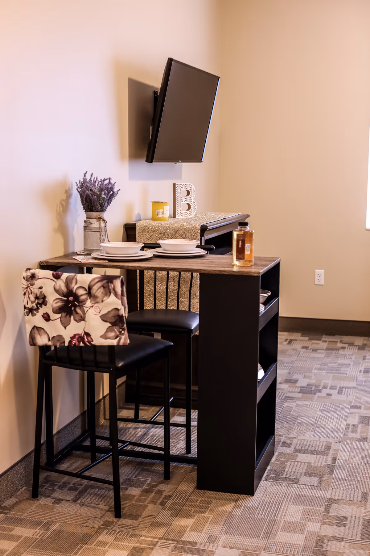 A small dining area with a high table set for two, featuring two chairs with floral patterned backs. On the table are two bowls and plates, a small yellow candle, and a bottle of liquid. Behind the table is a piano with a decorative letter B and a vase with purple flowers. A flat-screen TV is mounted on the wall above the piano. The room has beige walls and patterned carpet flooring.