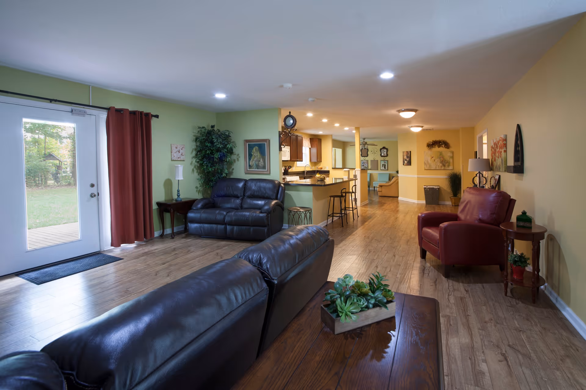 Interior view of a senior living facility common area with wooden flooring, leather sofas, a red armchair, a wooden coffee table with a plant centerpiece, and a kitchen area with bar stools in the background. The walls are painted green and yellow, and there is a glass door with red curtains leading outside.