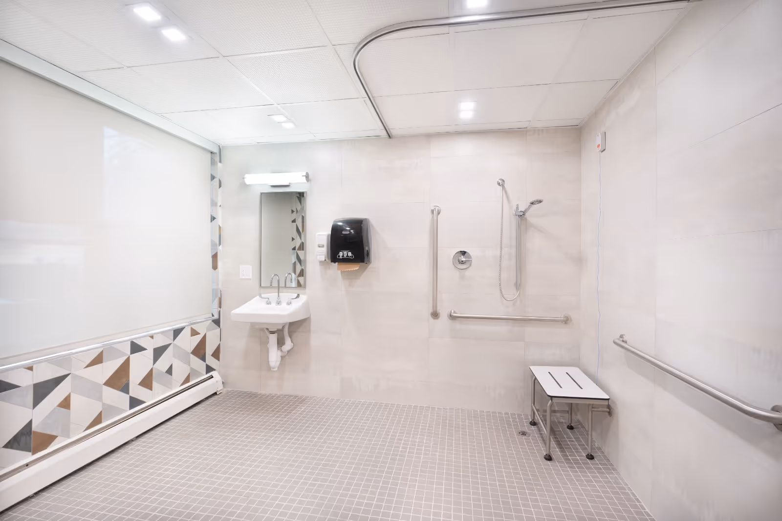 Accessible tiled bathroom with a wall-mounted sink and mirror, grab bars, handheld shower, and a folding shower bench.