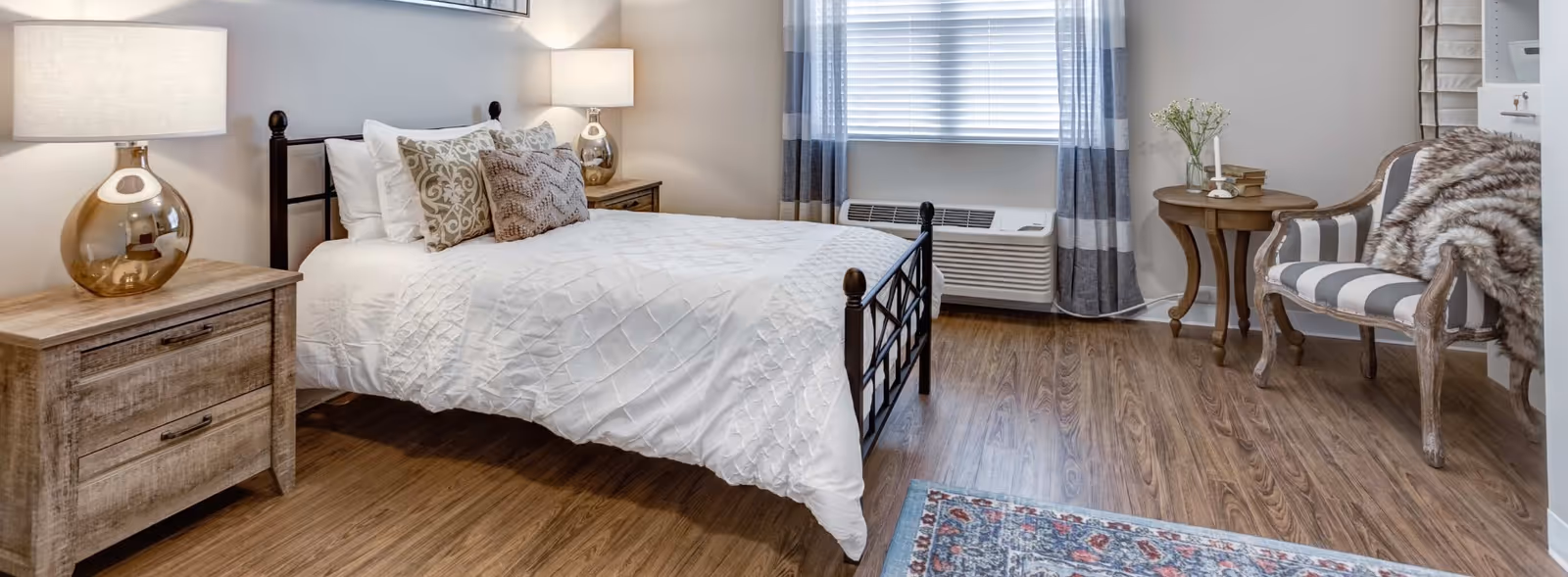A cozy bedroom with a wooden floor, featuring a bed with white bedding and decorative pillows. There are two wooden nightstands with lamps on either side of the bed. A window with striped curtains is behind the bed, and a striped armchair with a fur throw is next to a small round wooden table with a vase and candle. A patterned rug is partially visible on the floor.
