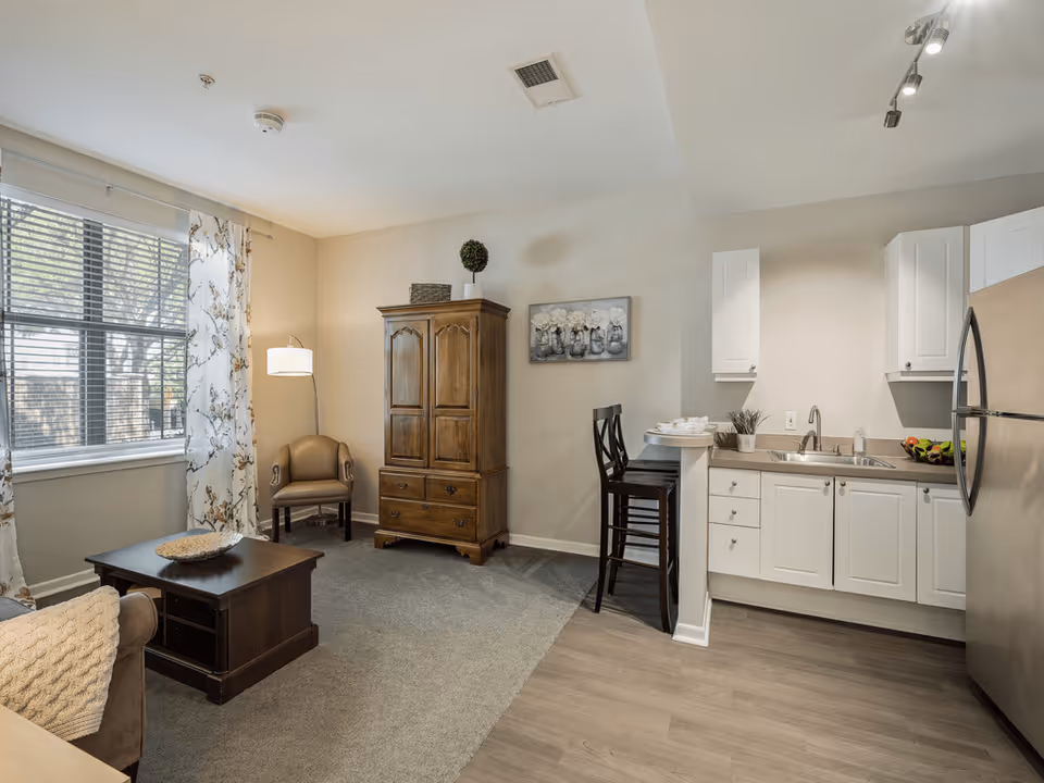 A cozy living area with a window covered by floral curtains, a wooden coffee table, a beige armchair, and a wooden armoire. Adjacent to the living area is a small kitchen with white cabinets, a stainless steel refrigerator, a sink, and a breakfast bar with two high chairs.
