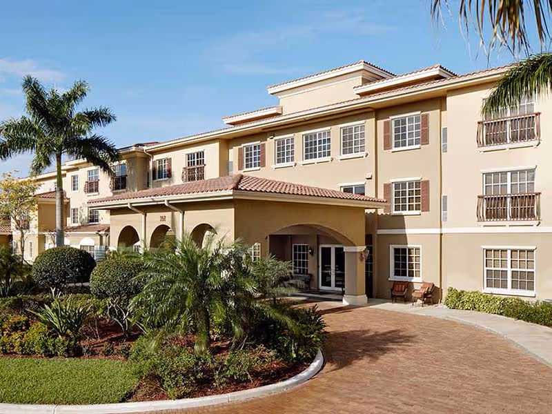 Exterior view of a three-story senior living facility building with beige walls and a red-tiled roof. The entrance features an arched covered driveway surrounded by well-maintained landscaping including palm trees and bushes under a clear blue sky.