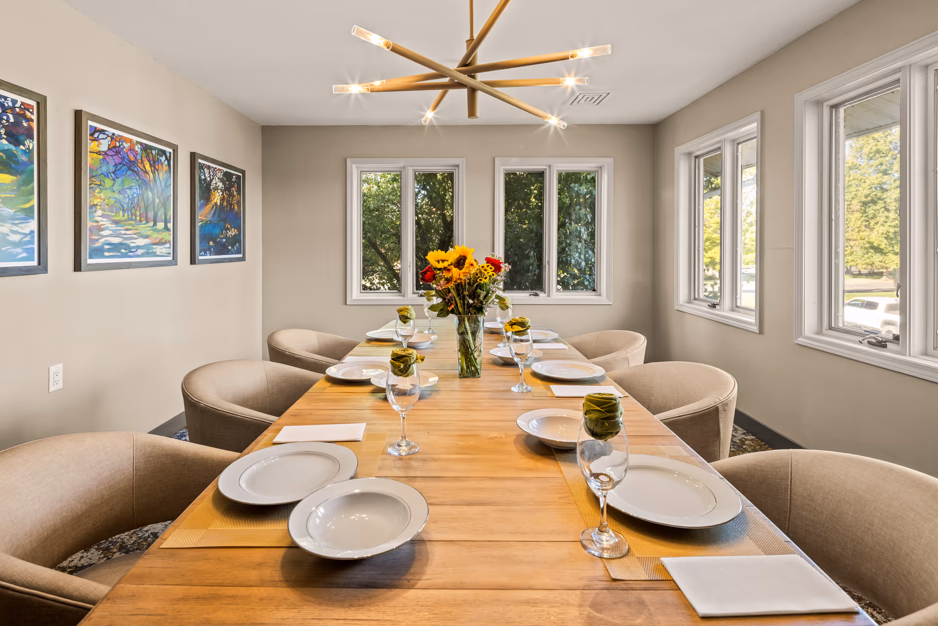 A bright dining room with a long wooden table set for eight people. Each place setting includes a white plate, a bowl, a wine glass with a green napkin inside, and a white napkin. A vase with sunflowers and other flowers is centered on the table. The room has beige walls, three colorful framed paintings on one wall, and multiple windows letting in natural light. A modern chandelier hangs above the table.