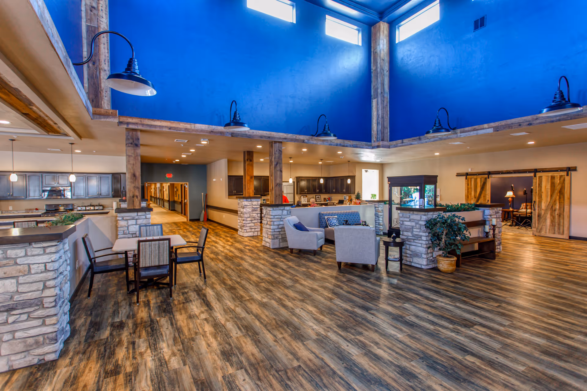 Spacious senior living facility common area with high blue walls and large windows near the ceiling. The room features wood-style flooring, stone half-walls, wooden beams, and modern light fixtures. There are seating areas with chairs and tables, a small aquarium, and a hallway leading to other rooms.