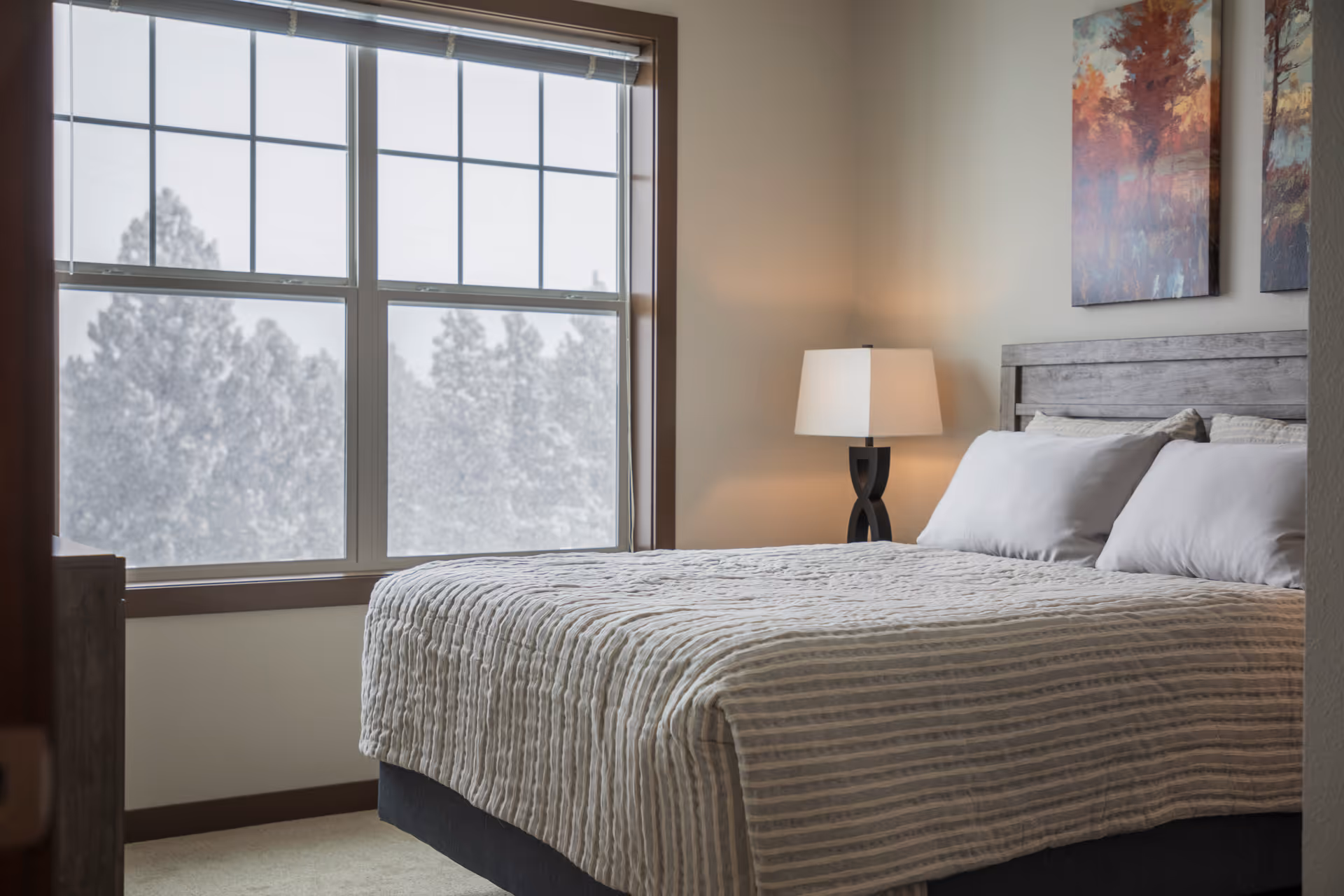 A cozy bedroom with a large window showing trees outside. The bed is made with a striped beige and white comforter and white pillows. There is a modern table lamp on a nightstand next to the bed, and two colorful abstract paintings hang on the wall above the headboard.