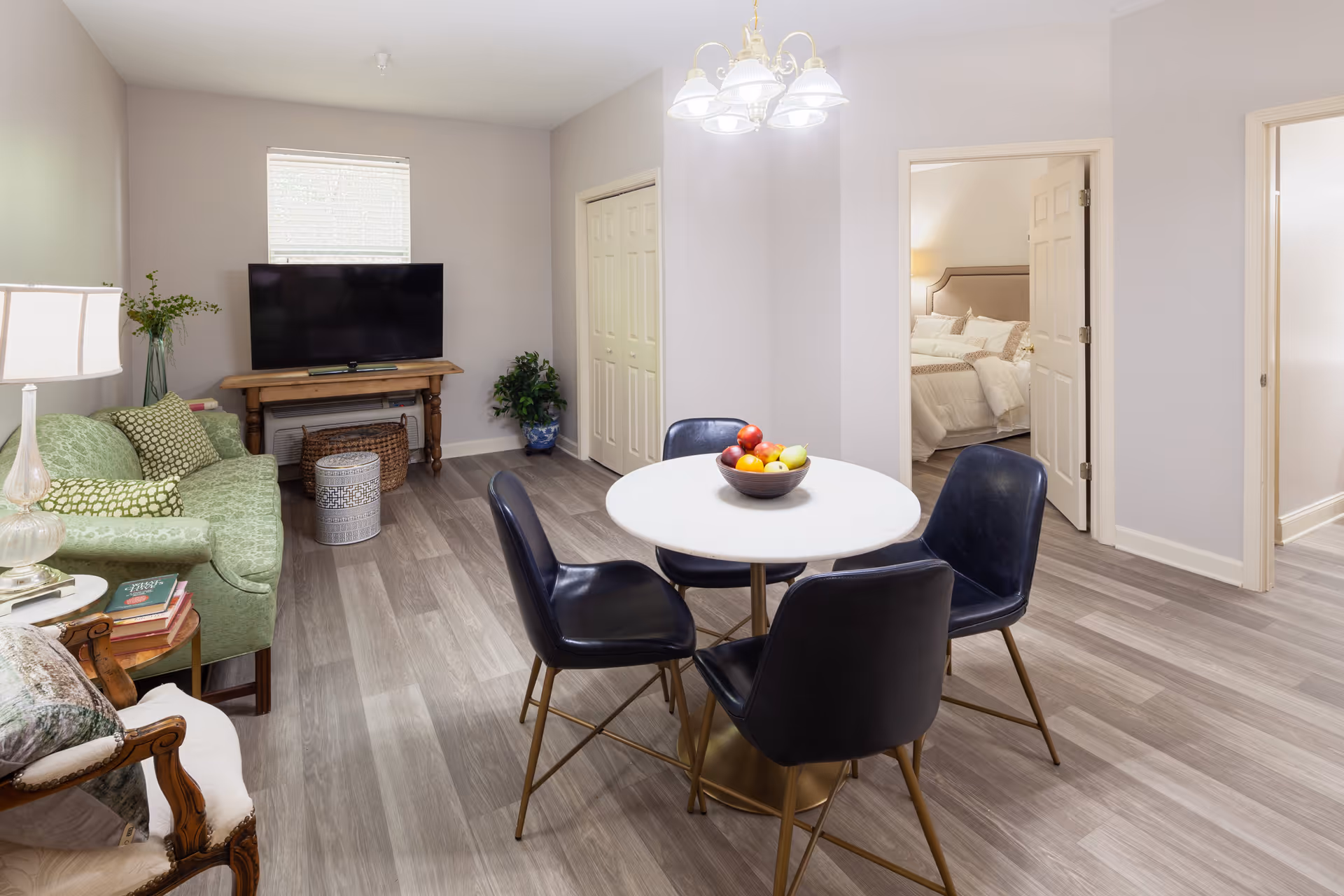 A cozy living room and dining area with a green couch, a wooden side table with books, a TV on a wooden stand, and a round white dining table with four black chairs. There is a bowl of fruit on the dining table. The room has light-colored walls and wood flooring. Two doorways lead to a bedroom with a bed and a hallway.