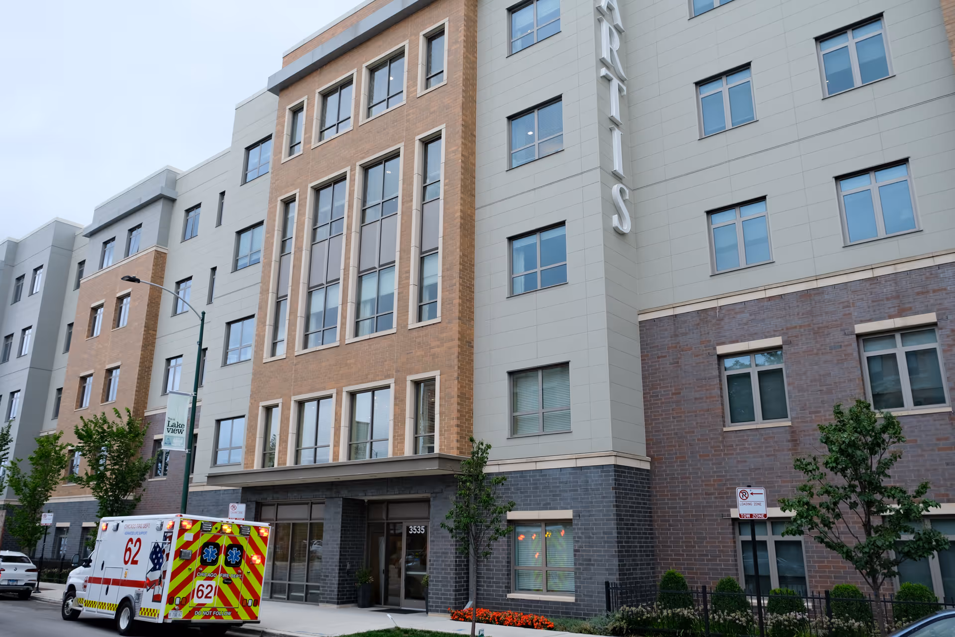 Exterior view of a multi-story senior living facility building with a mix of brick and light-colored paneling. There are several windows and a main entrance with the address number 3535. An ambulance is parked on the street in front of the building. Small trees and landscaping are visible along the sidewalk.