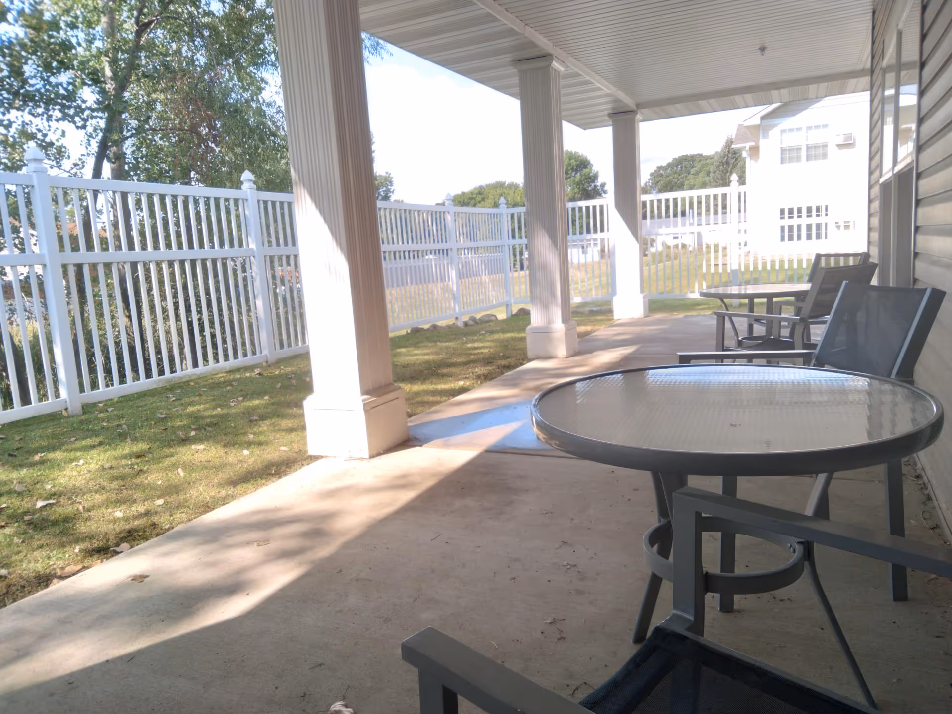Covered outdoor patio area with a glass-top round table and several chairs. The patio is supported by white columns and overlooks a grassy area enclosed by a white fence. Trees and a building are visible in the background.