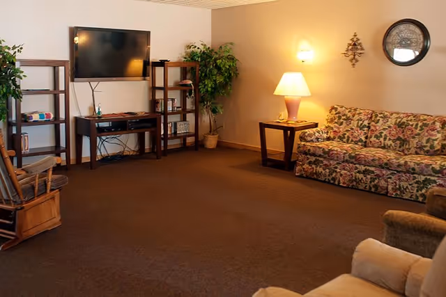 A cozy living room with a floral patterned sofa, a wooden rocking chair, a TV mounted on the wall with a wooden TV stand and shelves, a potted plant, a side table with a lamp, and a round decorative wall mirror.