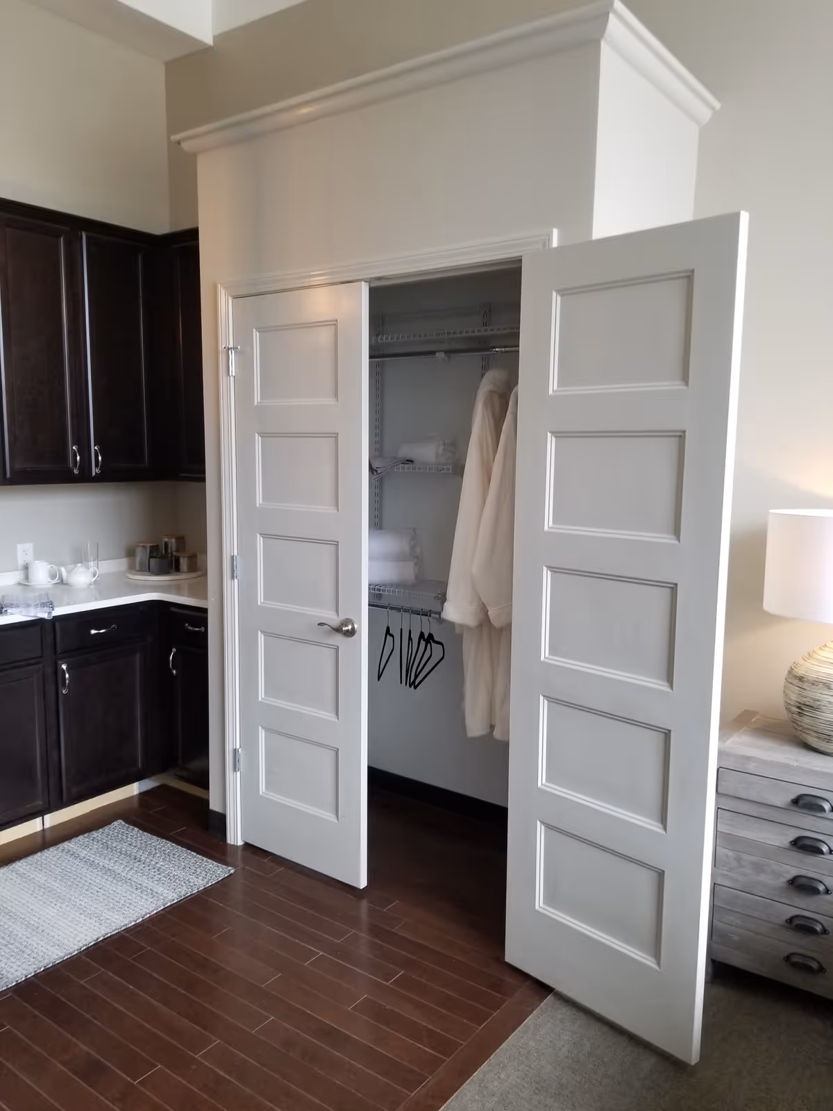 Interior view of a room featuring an open white double-door closet with hanging robes and folded towels inside. To the left, there are dark wood cabinets with a countertop holding a teapot and cups. To the right, a wooden nightstand with a lamp is partially visible. The floor is a combination of dark wood and carpet.