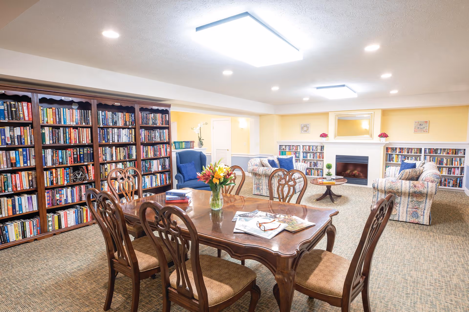 Well-lit communal library and lounge with bookshelves, a wooden table and chairs, upholstered sofas and armchairs, and a fireplace.