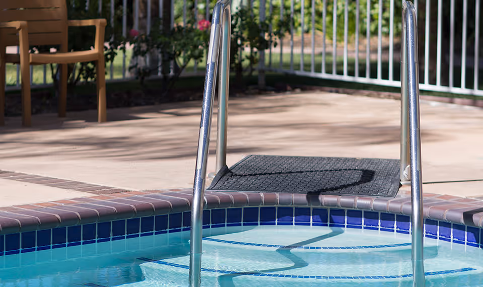 Close-up view of a pool ladder leading into a small pool or hot tub with blue and white tiled edges, a concrete pool deck, a brown chair, and a white metal fence in the background.