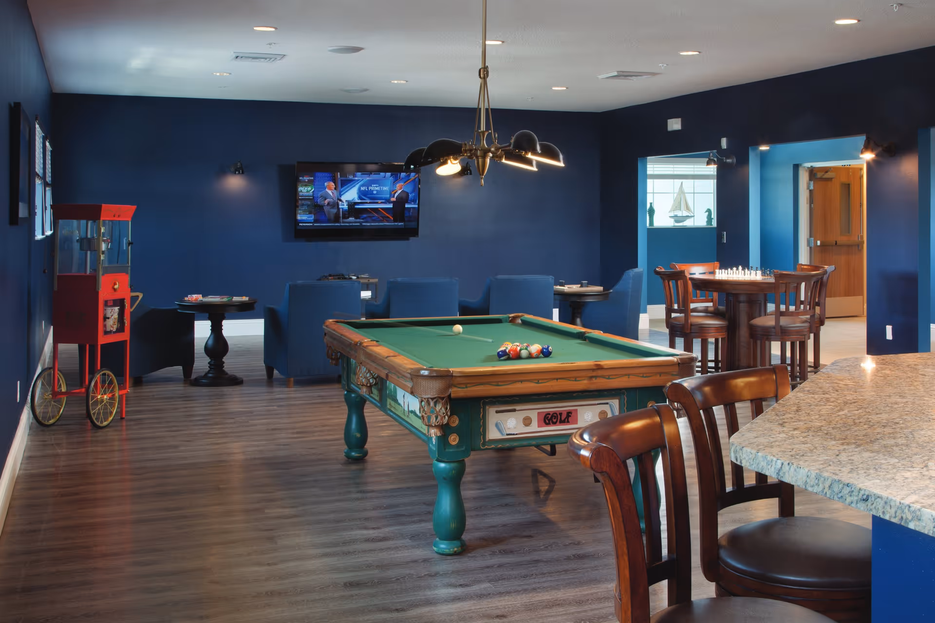 A blue-walled communal lounge with a pool table, seating area, wall-mounted TV, popcorn machine, and bar seating.