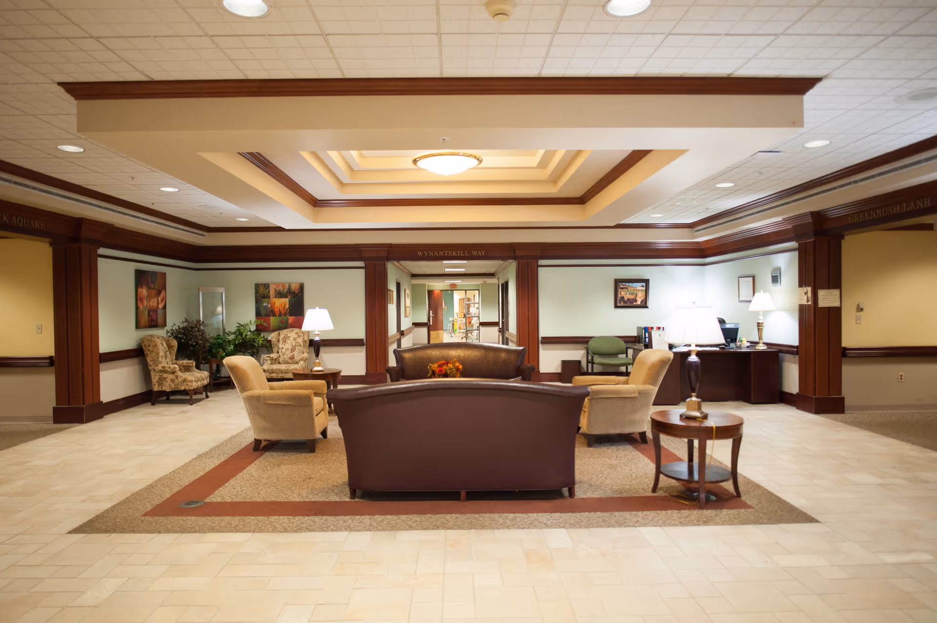 A spacious and well-lit common area in a senior living facility featuring a central seating arrangement with a brown leather sofa, two beige armchairs, and a round wooden side table with a lamp. The room has a decorative ceiling with recessed lighting, framed artwork on the walls, and additional seating with patterned armchairs and plants in the background. The area appears welcoming and comfortable with a mix of carpet and tile flooring.