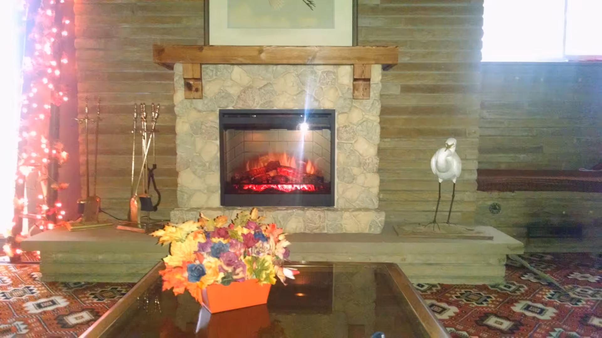 Cozy living room area featuring a stone fireplace with a wooden mantle, a glass coffee table with a colorful floral arrangement in an orange container, a decorative white bird sculpture on the right side of the hearth, fireplace tools on the left, and a patterned carpet on the floor. There are string lights with red leaves on the left side near a window.