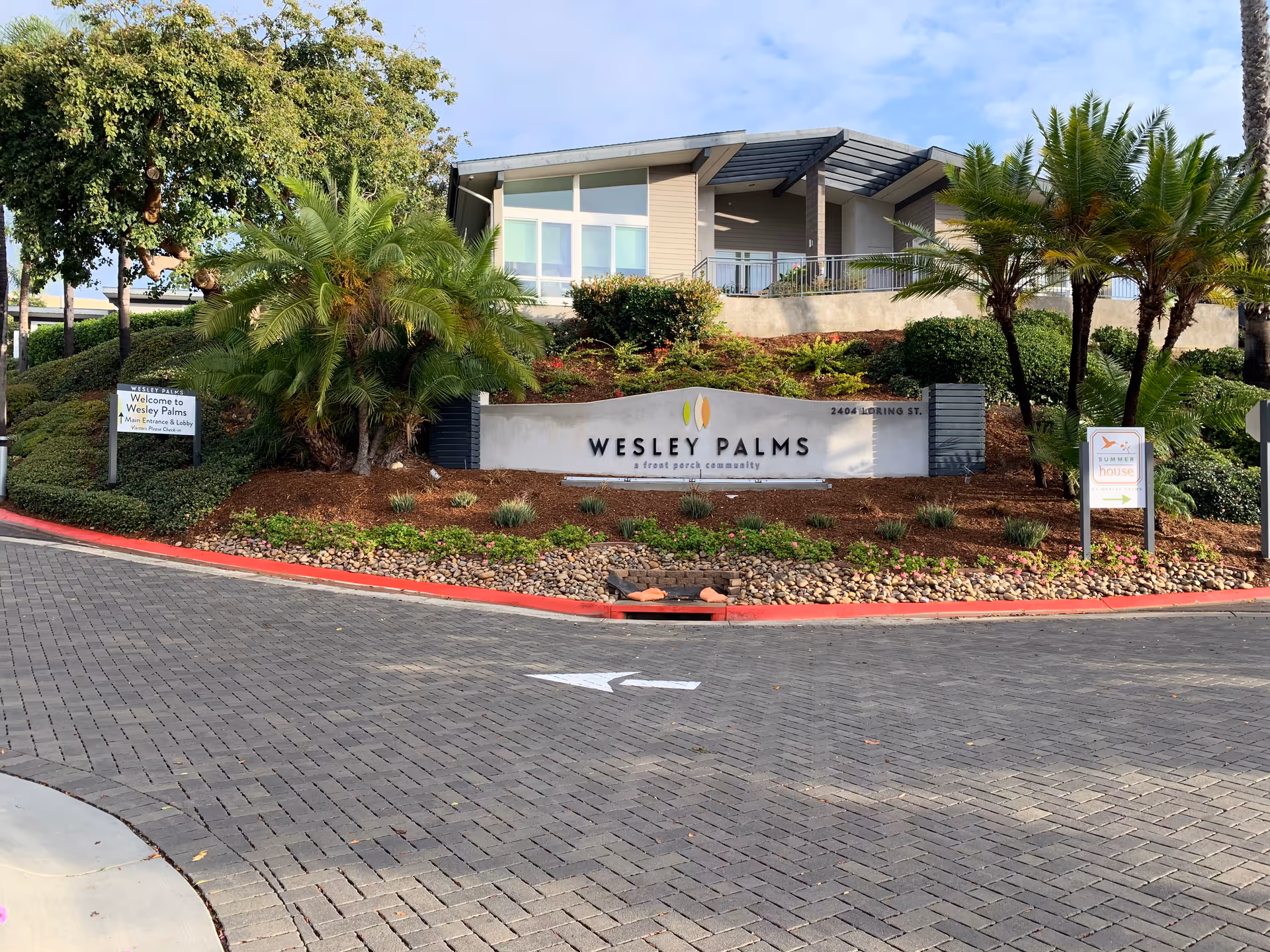 Entrance view of Wesley Palms Retirement Community showing a landscaped area with palm trees and bushes, a large sign with the community name, and a modern building in the background under a clear sky.