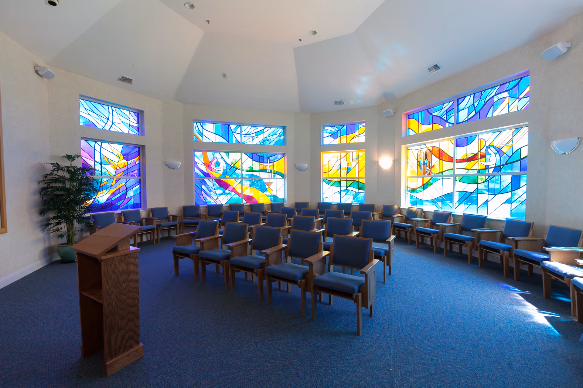 Interior of a chapel or meditation room with rows of wooden chairs with blue cushions arranged facing a wooden podium. The room features large colorful stained glass windows with abstract designs, beige walls, and blue carpeted floor.
