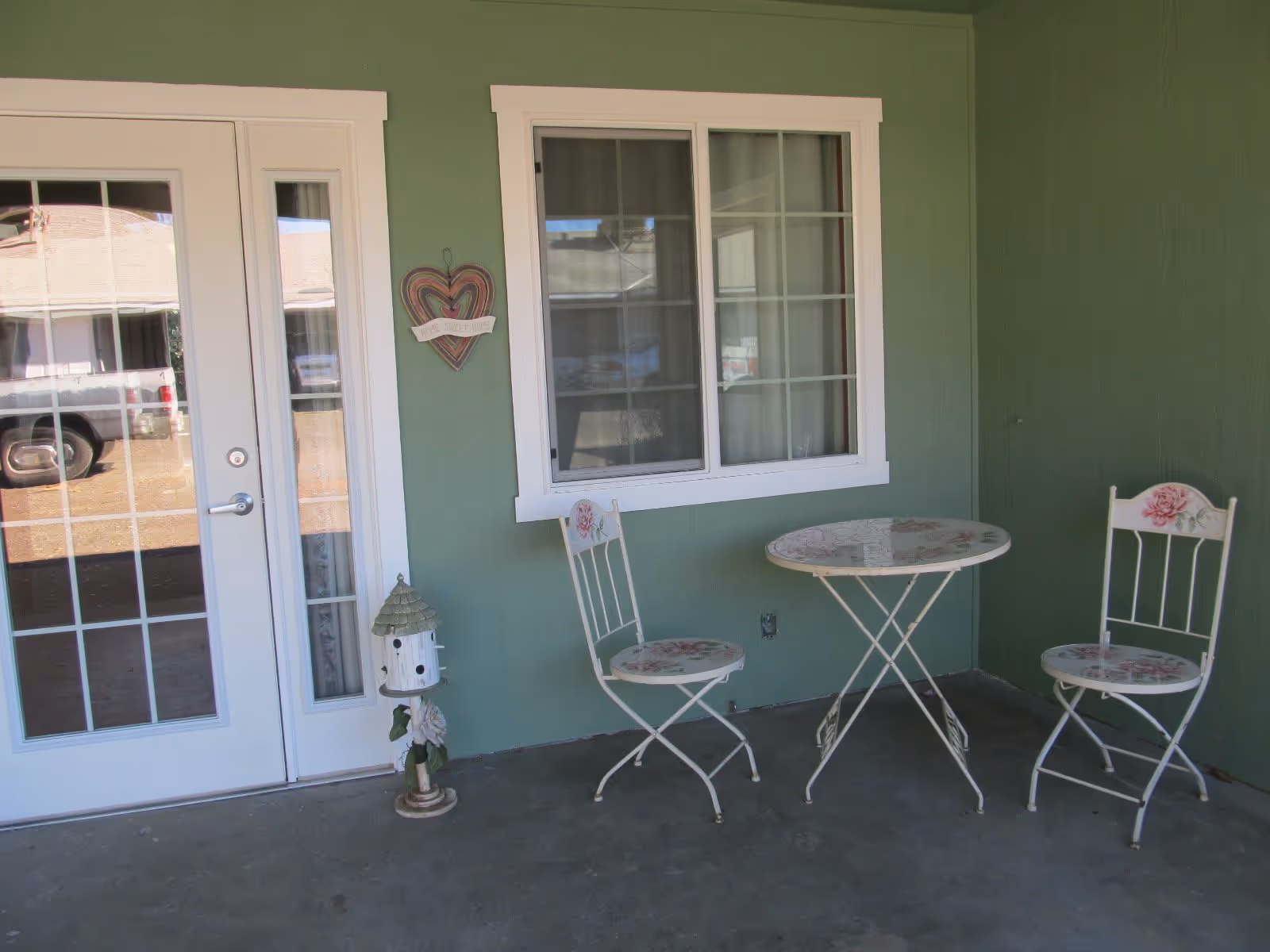A small outdoor patio area with green walls, a white-framed glass door, and a window. There is a small round table with floral patterns and two matching chairs. A decorative birdhouse and a heart-shaped wall hanging with the words 'Home Sweet Home' are also visible.