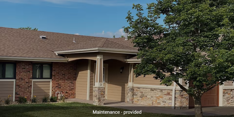 Exterior view of a single-story building with a brown roof, beige siding, and stone accents. There is a covered entrance with the number 2 on a pillar, a tree with green leaves on the right side, and a well-maintained lawn in front.