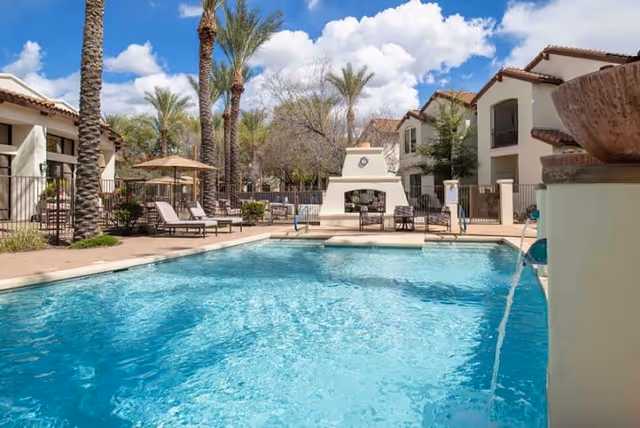 Outdoor swimming pool area with clear blue water, surrounded by lounge chairs, palm trees, and a white outdoor fireplace. Residential buildings with balconies are visible in the background under a partly cloudy sky.