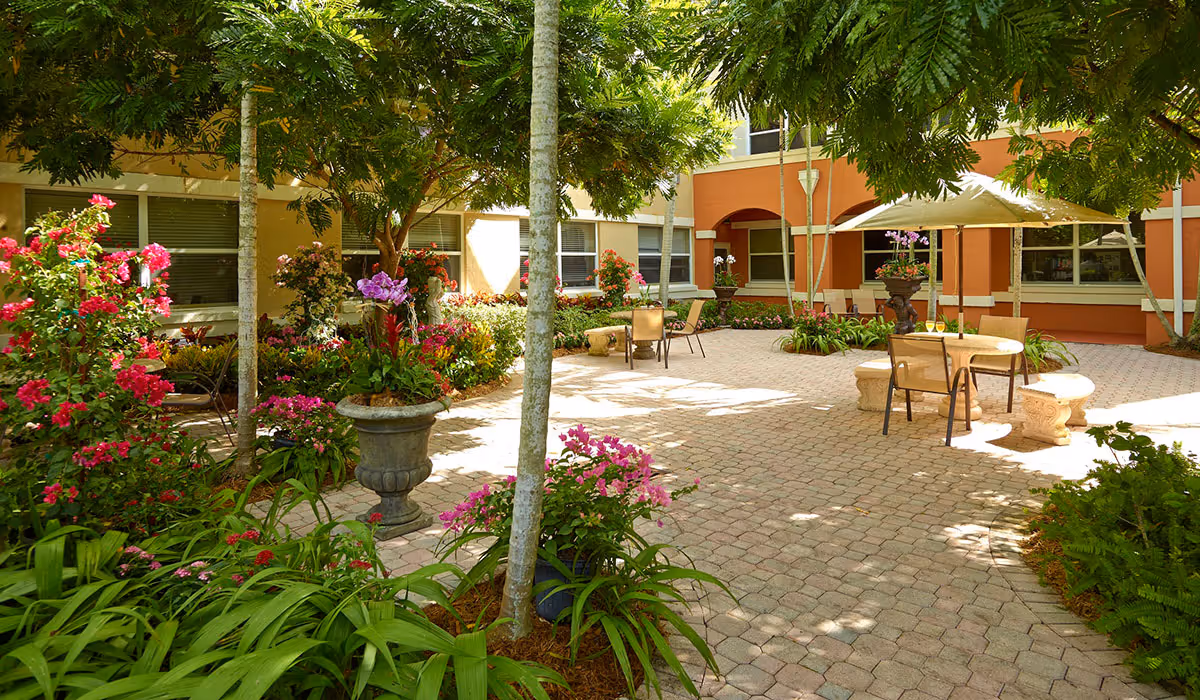 A sunny outdoor courtyard at The Palace Nursing and Rehabilitation Center featuring a paved patio with tables, chairs, and umbrellas surrounded by lush greenery, colorful flowers, and trees providing shade.