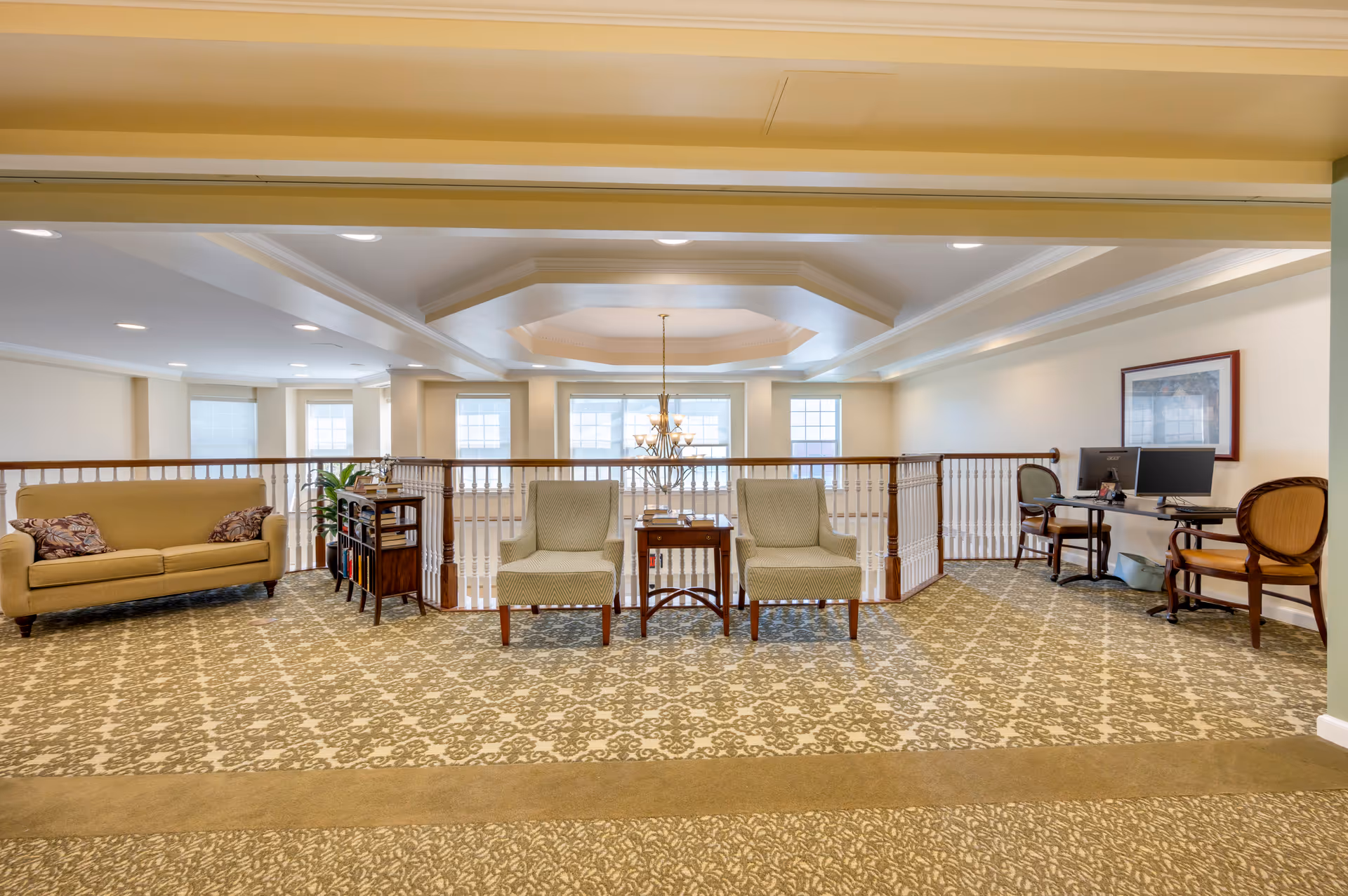 A spacious, well-lit common area with patterned carpet flooring, featuring a beige sofa with two decorative pillows on the left, two cushioned chairs with a small wooden table between them in the center, and a desk with two computer monitors and chairs on the right. The area overlooks a railing with large windows and a chandelier visible in the background.