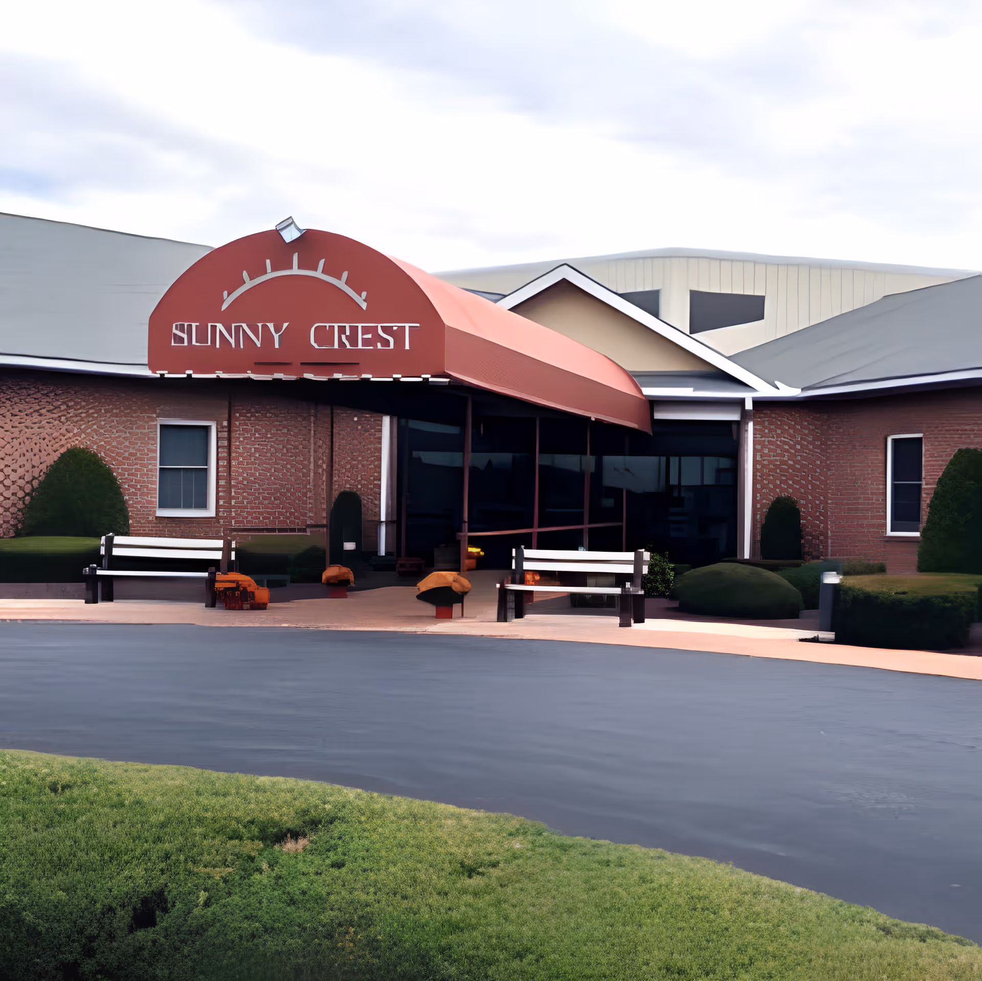 Front exterior view of Sunny Crest Home, Inc with a red awning over the entrance, two white benches on either side of the walkway, and neatly trimmed bushes and grass in the foreground.