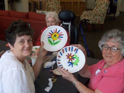 Three elderly women sitting around a table in a common area, holding up paper plates decorated with colorful flower designs. One woman is in a wheelchair, and the room has floral-patterned armchairs and a piano in the background.