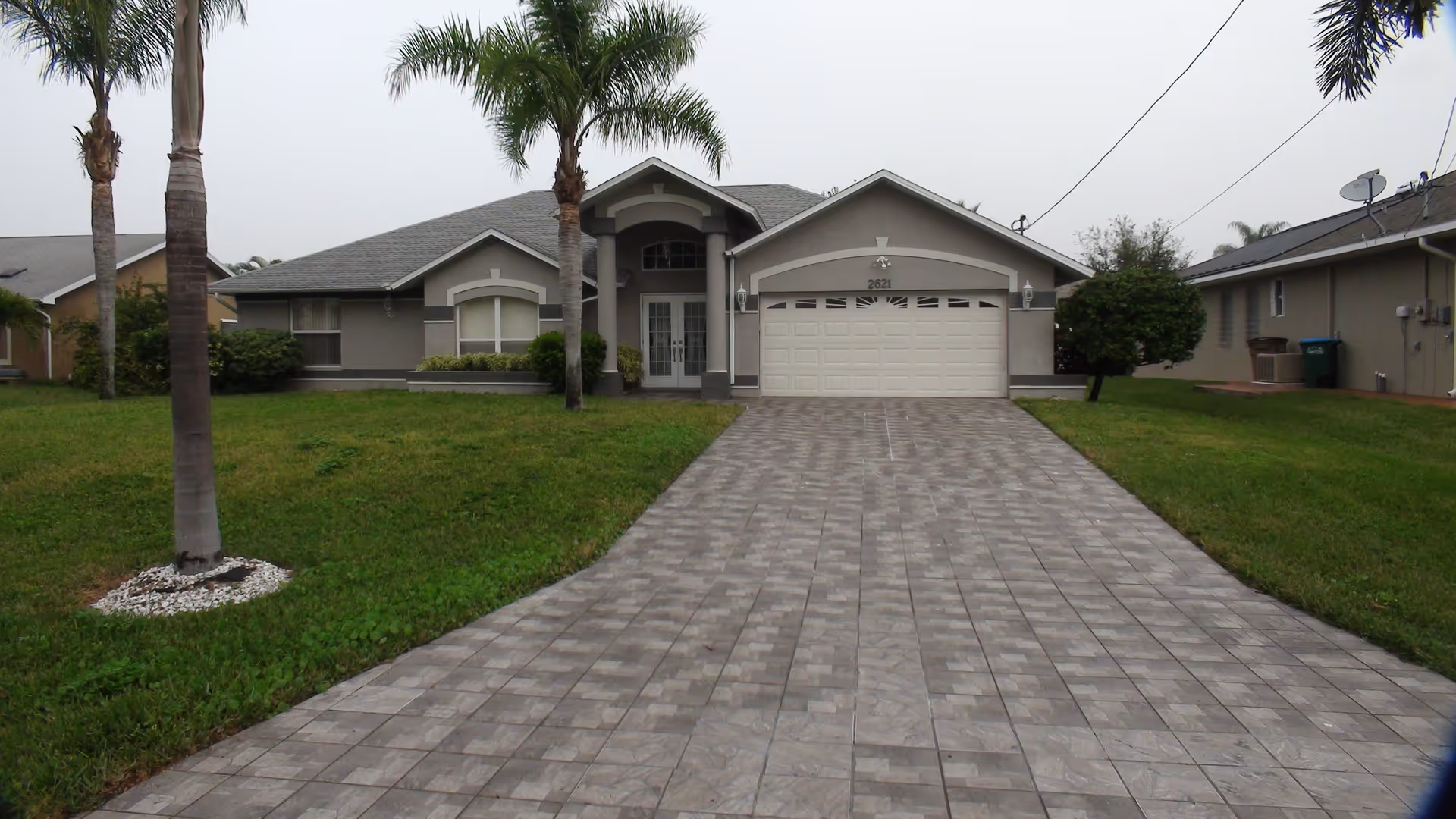 Front exterior view of a single-story residential building with a tiled driveway, two palm trees on the lawn, and a two-car garage door under a gabled roof.