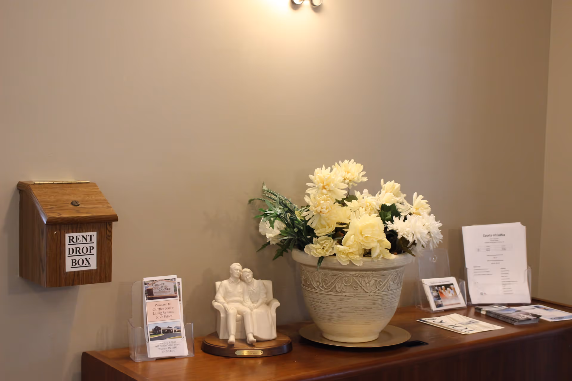 A wooden rent drop box mounted on a beige wall above a wooden surface displaying a white ceramic figurine of an elderly couple sitting on a bench, a large decorative pot with cream-colored flowers, and various brochures and papers including one titled 'Courts of Colfax'.